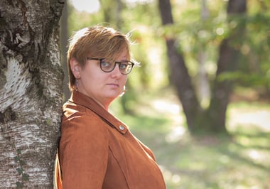 Femme avec lunettes et veste marron adossée à un arbre en forêt