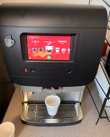 Super-automatic coffee machine dispensing a cappuccino into a paper cup with touchscreen interface.