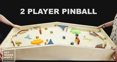 2 people standing in front of a 2-player pinball game table. 