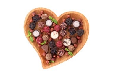A chocolate filled wooden, heart shaped bowl on a white background. Mint leaves, hazelnuts, truffles