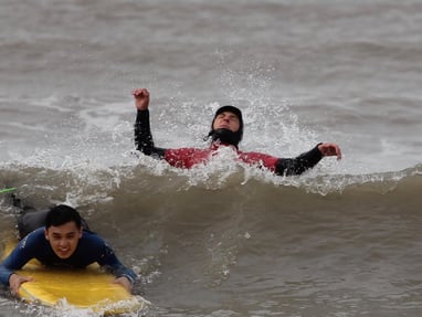 A prone smiling surfer dropping down a wave while a surf instructor jumps backwards over a wave.