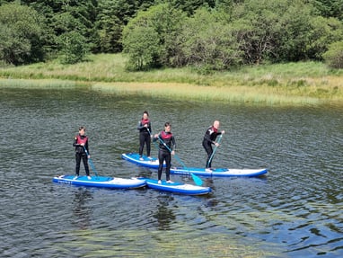 A group of four stand up paddle boarders in wetsuits and buoyancy aids on a calm lake.