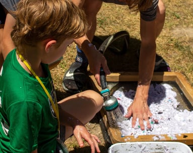 Teacher using a hose to spray shredded paper onto a screen with kids surrounding.