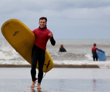 A stoked surf instructor carrying a yellow surfboard, giving a shaka and sticking his tounge out.