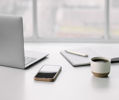 a laptop, cell phone, notebook with pen, and a cup of coffee on a desk
