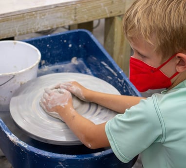 Young child wearing a mask with both hands covering a mound of clay on a pottery wheel.