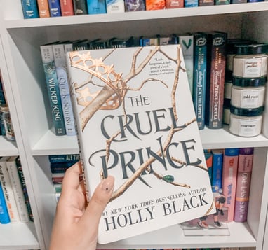 Mariah is holding up a hardcover copy of The Cruel Prince by Holly Black in front of her shelves.