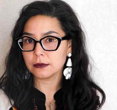 Felicia Rose Chavez looks at the camera wearing thick, angular glasses, large white earrings.