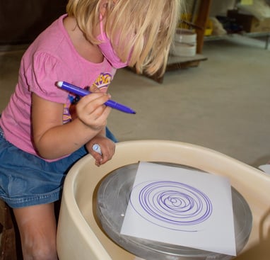 Young girl sitting at a potters wheel holding a marker, making spin art.