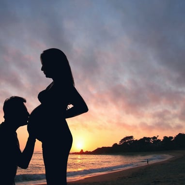 Um casal, sendo ela, grávida, e ele beijando sua barriga, numa praia deserta, ao por do sol.
