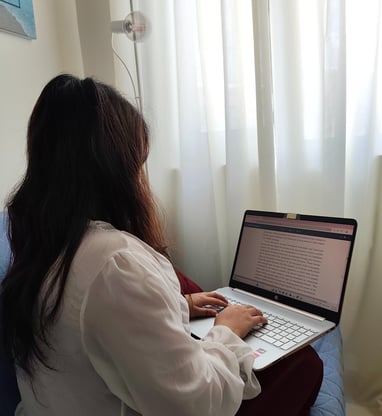 Woman typing on a laptop with a document open, sitting near a window with white curtains.