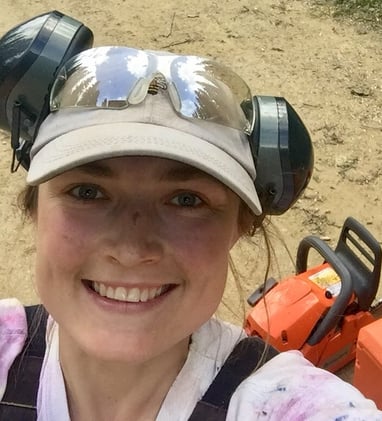 Keely smiles wide, wearing a beige ballcap and headphones. Behind her is an orange chainsaw.