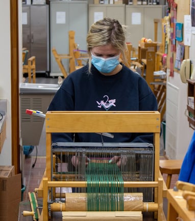 A young woman standing in front of a table loom adjusting your piece.