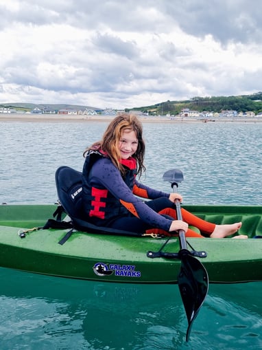 A girl kayaking in a sit on top kayak in the sea with Borth village and cloudy skies behind her