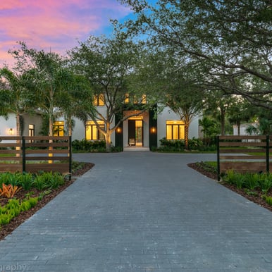 a brick driveway leading up to a well lit luxury home at sunset time with a vibrantly lit sky