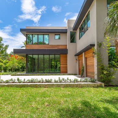 front yard of a luxury square looking home with a driveway, two garages and a forest. 