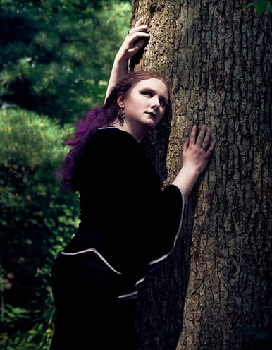 a woman with purple hair in a black dress in the forest with sunlight on her face