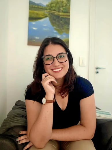Psicóloga Rafaela Antoniazzi sorrindo, vestindo óculos e blusa preta, apoiando o rosto na mão. Seção Sobre.