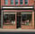 A charming bakery storefront with a brick and pastel blue facade. The lettering on the sign reads 'Main Street Bakery', adorned with small decorative bulbs. The awning is a bright blue, complementing the calm and inviting exterior. Next door, another shop offers fresh goods like bread, cookies, and cakes, as indicated by the signage.