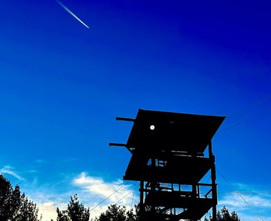 Silhouette of a fire tower against a deep blue sky with a jet trail overhead, surrounded by treetops and clouds.
