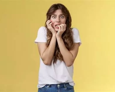 Femme cheveux longs apeurée yeux exorbités, main droite sur joue, doigts main gauche dans la bouche