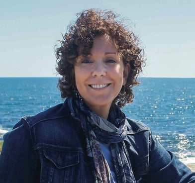 Aggie, with short curly hair and a denim jacket, stands in front of a bright blue sea.