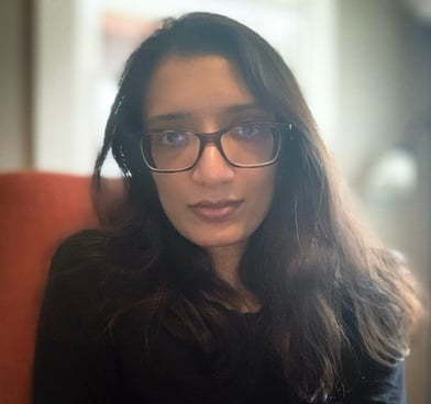 Shreya Fadia wears large square glasses and a black shirt. Her dark hair is long and wavy.