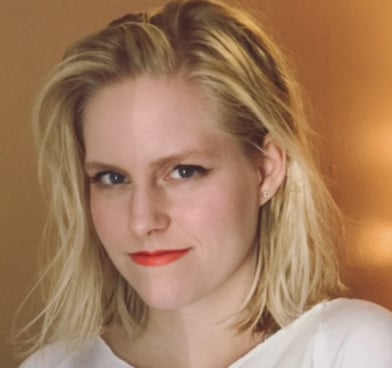 Ellie Black smiles at the camera, wearing a white shirt and standing in front of an orange wall.