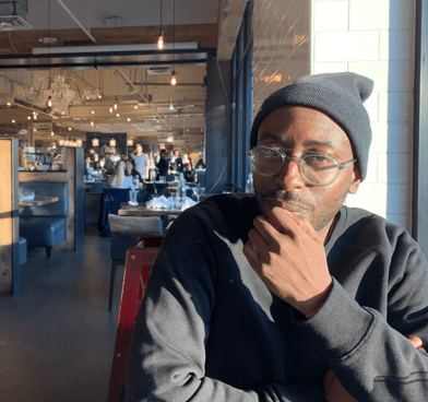 Koyé Oyedeji wears glasses, a beanie, and a black sweatshirt. His hand rests near his chin.