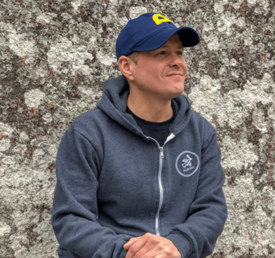 Alejandro Puyana wears a blue cap and a gray zip-up sweatshirt. He looks beyond the camera, smiling.