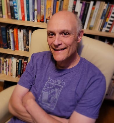 Gregory Spatz sits in an armchair in front of a bookshelf. He wears a purple shirt and smiles.