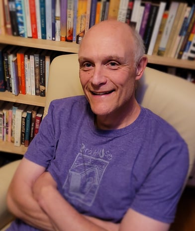 Gregory Spatz sits in an armchair in front of a bookshelf. He wears a purple shirt and smiles.