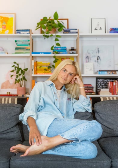 Natalie Warther sits on a grey couch in front of books and plants. She is blonde and wears denim.