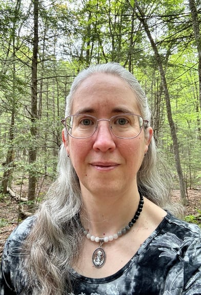 Portrait of a middle-aged white woman with long gray hair and glasses, standing in the woods.