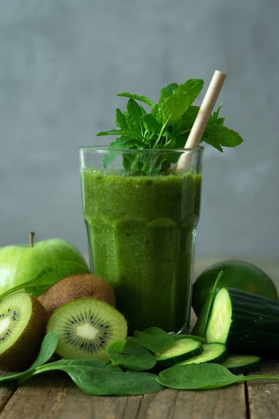 Copo de suco verde com folhas de hortelã, rodeado de ingredientes naturais como kiwi, pepino, maçã