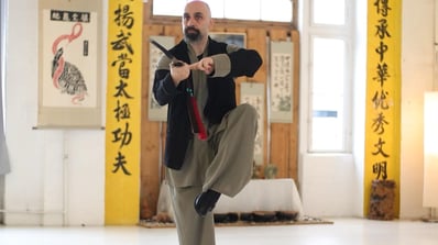 Ismet Himmet demonstrating a balanced Wudang sword (Jian) form indoors, with Daoist scrolls behind.