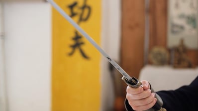 Close-up of a hand holding a Wudang Jian sword, with engraved blade and yellow calligraphy scroll behind.