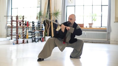 Ismet Himmet practicing a Wudang Jian sword form indoors, in a deep stance near weapon racks.