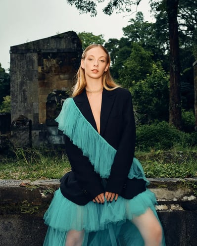 Portrait of model in a blue ruffle suit standing in front of a ruin in a forest, styled for a fashion editorial.