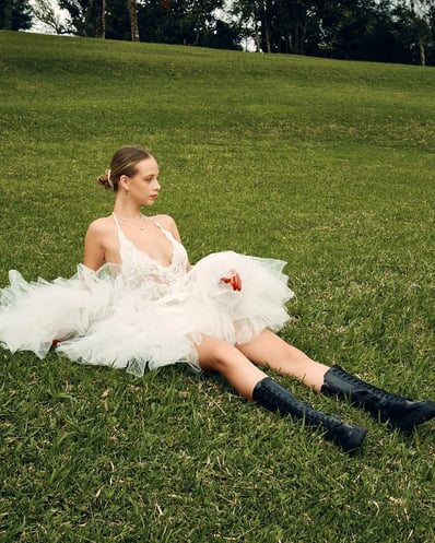 Full-body shot of model in a white lace top sitting on grass in a garden, styled for a fashion editorial.