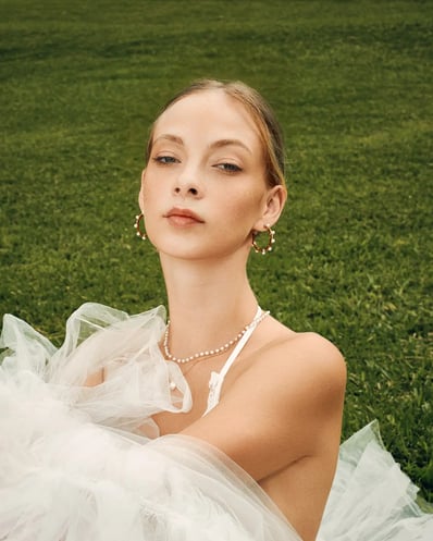 Portrait of model in a white lace top sitting on grass in a garden, styled for a fashion editorial.