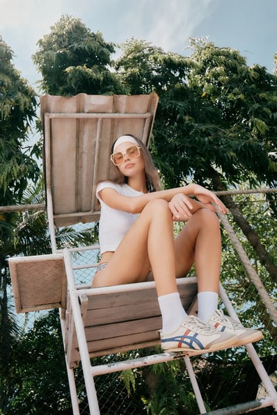 Model relaxes on an umpire chair wearing retro sunglasses and a white top, with soft outdoor tones.