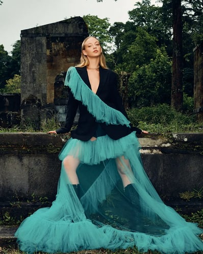 Full-body shot of model in a blue ruffle suit standing in front of a ruin in a forest, styled for a fashion editorial.