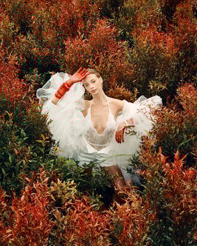 Full-body shot of model in a white lace top standing amid red flowers in a Bali garden, fashion editorial style.