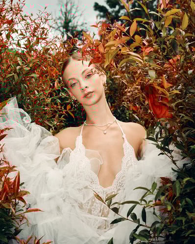 Portrait of model in a white lace top, framed by red flowers in a natural setting for a fashion editorial.