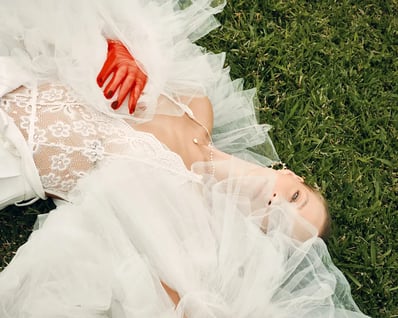 Portrait of model in a white lace top lying on grass with a tulle shawl partially covering her face, styled for an editorial.