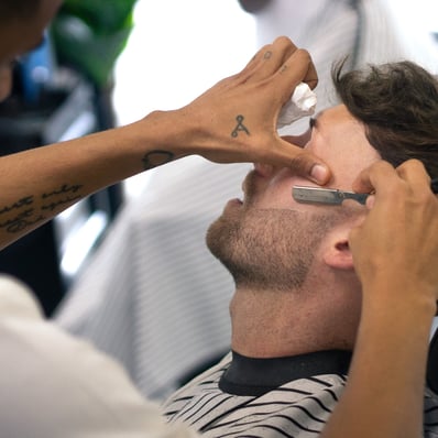 man receiving a shave with a cut throat razor by a barber while sitting in a chain in a barbershop