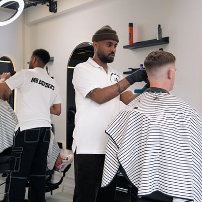 two barbers tending to cutting the hair of two customers while they are sitting in the chair