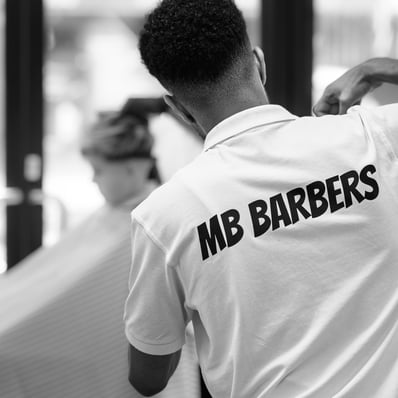 The back of a barber cutting a customers hair, with a blurred customer in the background