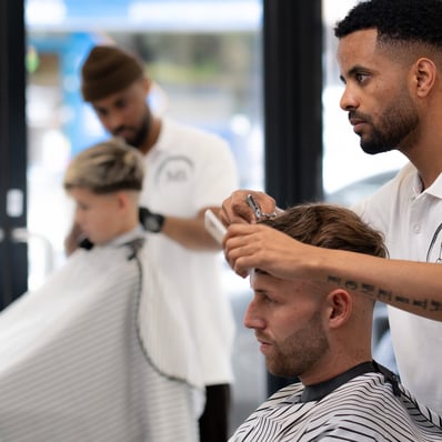two barbers tending to cutting the hair of two customers while they are sitting in the chair